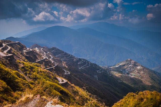 A wide-angle view of a long, winding mountain road zig-zagging up steep, grassy hills under a cloudy blue sky.