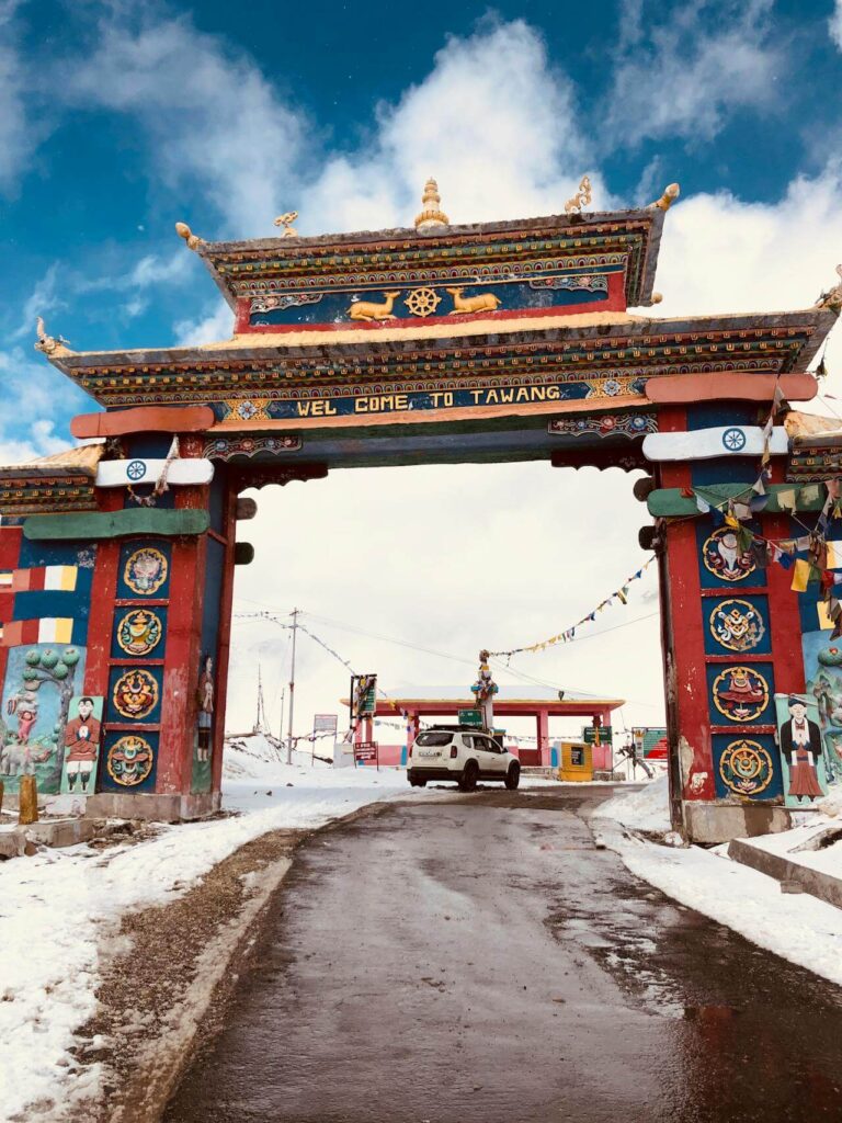 A colorful Buddhist-style welcome gate stands over a snowy mountain road, with the text "WELCOME TO TAWANG" inscribed across the top. The ornate structure is decorated with traditional motifs and prayer flags, set against a bright blue sky with white clouds.